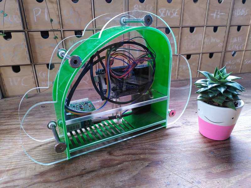 Photo of clear cloud shaped housing with electronics inside. There is a succulent in a pot with a smiley face on it next to it, and lots of small wood component drawers behind.