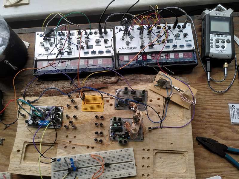 A slice of wood with circuits screwed to the top, connected by wires to two small volca modular synthesisers and a zoom recorder on a wooden workbench