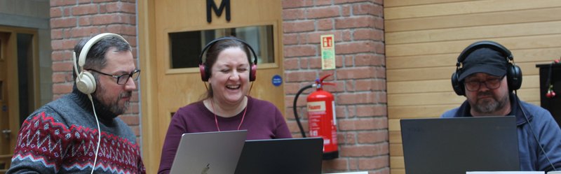 photo of three people playing the game at laptops, with headphones on, the person in the middle has a beaming smile and laugh
