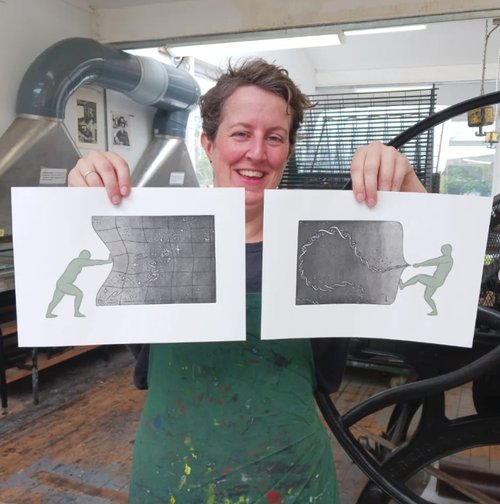 Photo of Amber in a print room next to a big iron press, holding up two prints and grinning