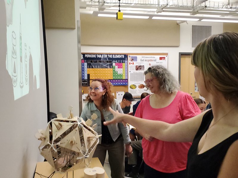Three adults looking animated around a wood virus structure