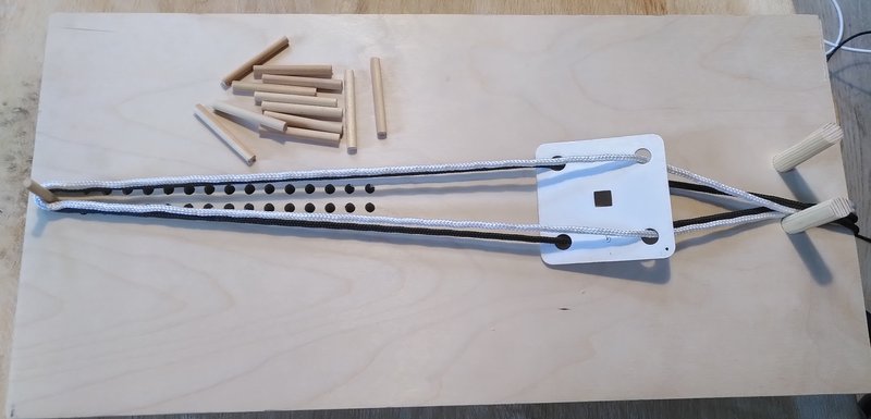 A wooden board with a tablet held by warp threads attached to a peg, a series of holes and a pile of pegs to put in them