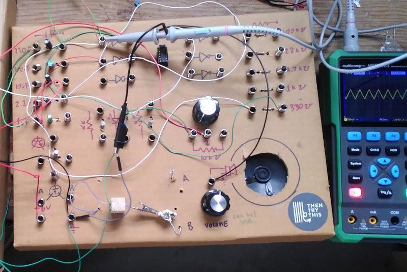 A cardboard box with springs and components with a small speaker inside on the left, wired up into a circuit. To the right of it an oscilloscope showing a triangle wave form.