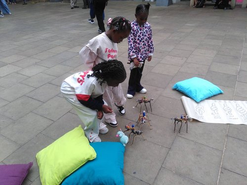 Three children playing with small woven robots with cushions scattered around.