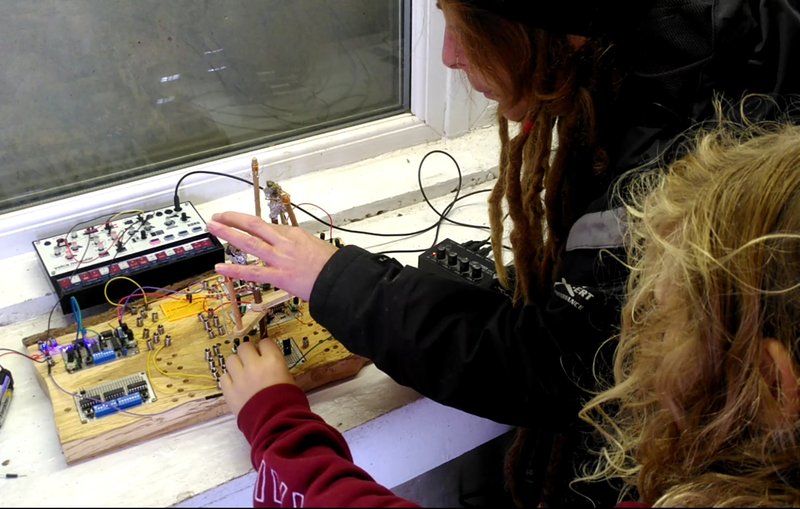 Two people operate a synth made from a chunk of natural wood and springs and wires